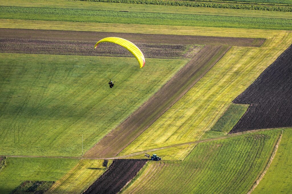 A paraglider enjoying a sunny day flying over expansive rural farmland.